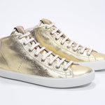 Three quarter front view of mid top gold sneaker with full leather upper with perforated crown logo, internal zip and white sole.