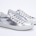 Three quarter front view of low top silver sneaker with perforated crown logo on upper. Full metallic leather upper and white rubber sole.