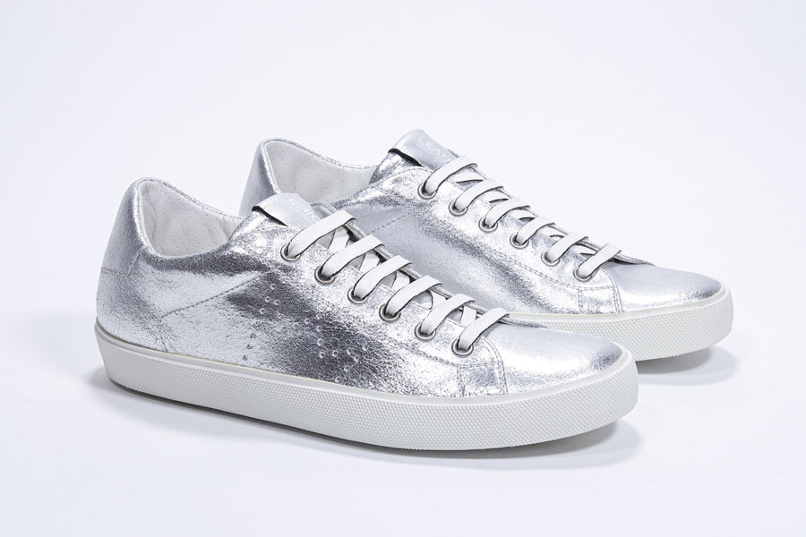 Three quarter front view of low top silver sneaker with perforated crown logo on upper. Full metallic leather upper and white rubber sole.