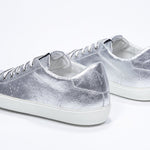 Three quarter back view of low top silver sneaker with perforated crown logo on upper. Full metallic leather upper and white rubber sole.