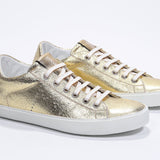 Three quarter front view of low top gold sneaker with perforated crown logo on upper. Full metallic leather upper and white rubber sole.