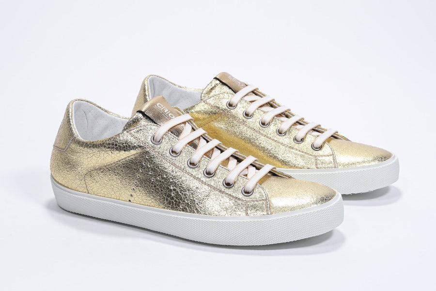 Three quarter front view of low top gold sneaker with perforated crown logo on upper. Full metallic leather upper and white rubber sole.