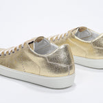 Three quarter back view of low top gold sneaker with perforated crown logo on upper. Full metallic leather upper and white rubber sole.