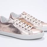 Three quarter front view of low top rose-gold sneaker with perforated crown logo on upper. Full metallic leather upper and white rubber sole.