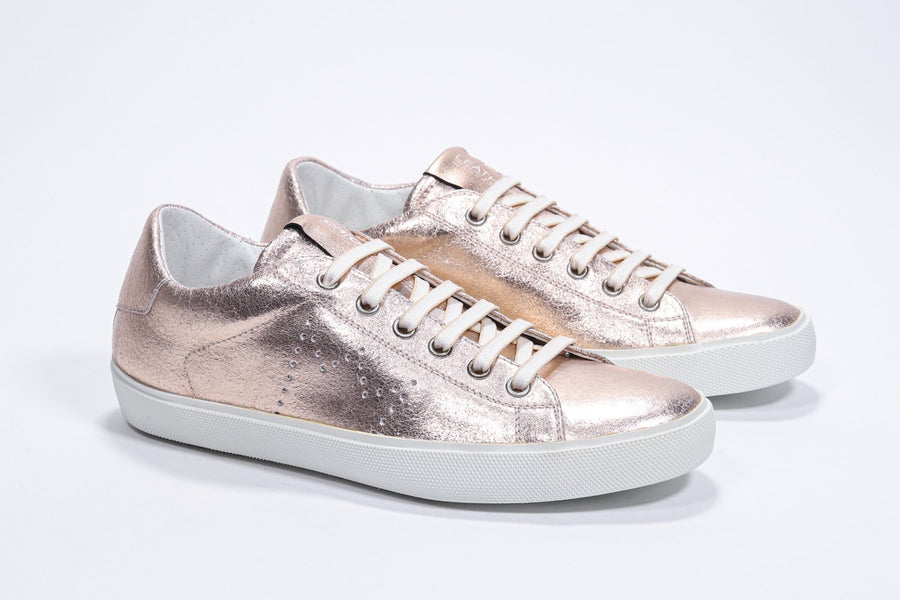 Three quarter front view of low top rose-gold sneaker with perforated crown logo on upper. Full metallic leather upper and white rubber sole.