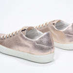 Three quarter back view of low top rose-gold sneaker with perforated crown logo on upper. Full metallic leather upper and white rubber sole.