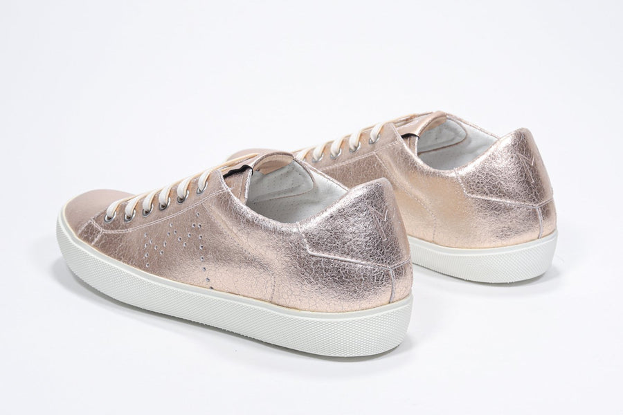 Three quarter back view of low top rose-gold sneaker with perforated crown logo on upper. Full metallic leather upper and white rubber sole.