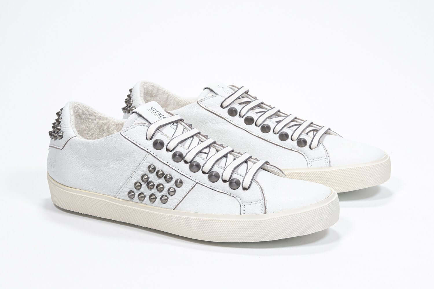 Three quarter front view of low top white sneaker. Full leather upper with studs and vintage rubber sole.