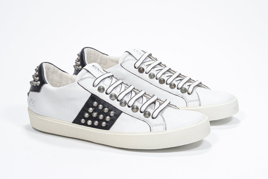 Three quarter front view of low top white and black sneaker. Full leather upper with studs and vintage rubber sole.