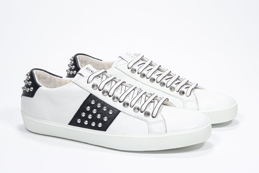 Three quarter front view of low top white and black sneaker. Full leather upper with studs and white rubber sole.