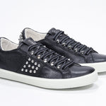Three quarter front view of low top black sneaker. Full leather upper with studs and white rubber sole.