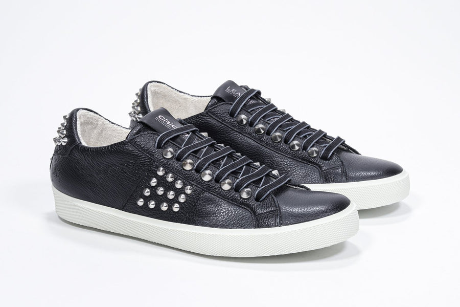 Three quarter front view of low top black sneaker. Full leather upper with studs and white rubber sole.