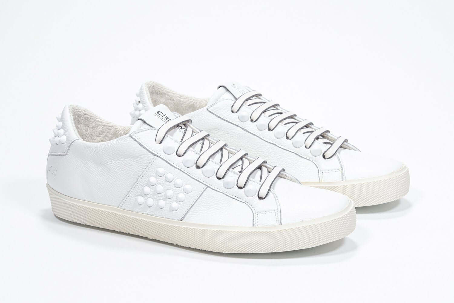 Three quarter front view of low top white sneaker. Full leather upper with studs and vintage rubber sole.
