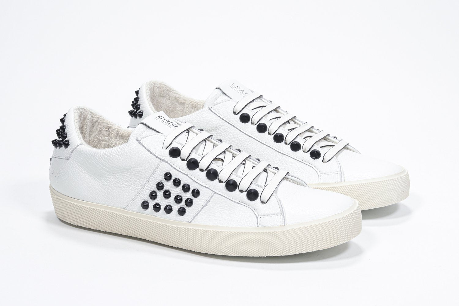 Three quarter front view of low top white sneaker. Full leather upper with studs and vintage rubber sole.