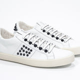 Three quarter front view of low top white sneaker. Full leather upper with studs and vintage rubber sole.