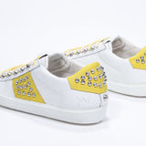 Three quarter back view of low top white and yellow sneaker. Full leather upper with studs and white rubber sole.