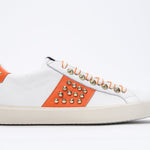 Side profile of low top white and orange sneaker. Full leather upper with studs and vintage rubber sole.