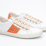 Three quarter front view of low top white and orange sneaker. Full leather upper with studs and vintage rubber sole.