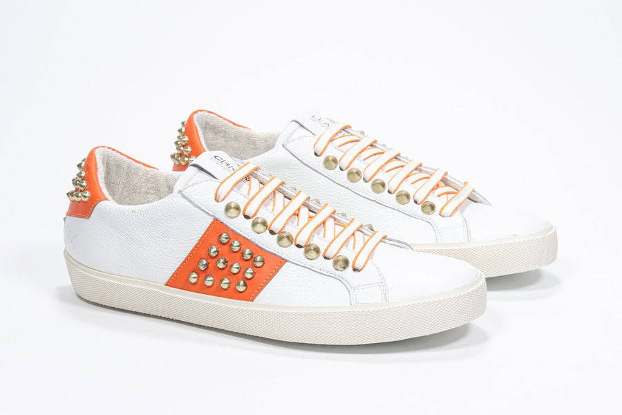 Three quarter front view of low top white and orange sneaker. Full leather upper with studs and vintage rubber sole.