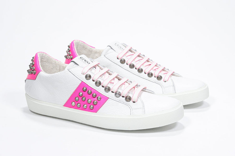 Three quarter front view of low top white and neon pink sneaker. Full leather upper with studs and white rubber sole.
