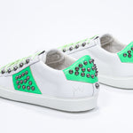 Three quarter back view of low top white and neon green sneaker. Full leather upper with studs and white rubber sole.