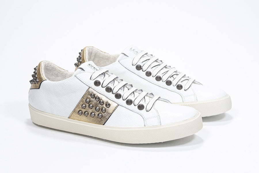 Three quarter front view of low top white and metallic gold sneaker. Full leather upper with studs and vintage rubber sole.