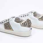 Three quarter back view of low top white and metallic gold sneaker. Full leather upper with studs and vintage rubber sole.