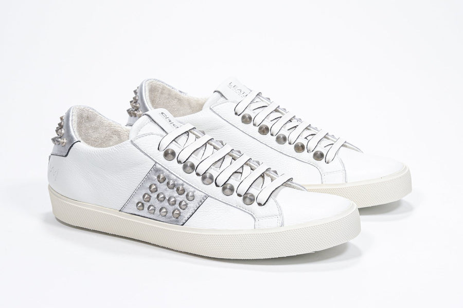 Three quarter front view of low top white and metallic silver sneaker. Full leather upper with studs and vintage rubber sole.
