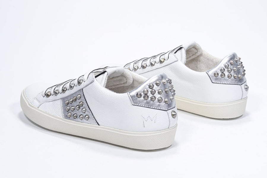 Three quarter back view of low top white and metallic silver sneaker. Full leather upper with studs and vintage rubber sole.