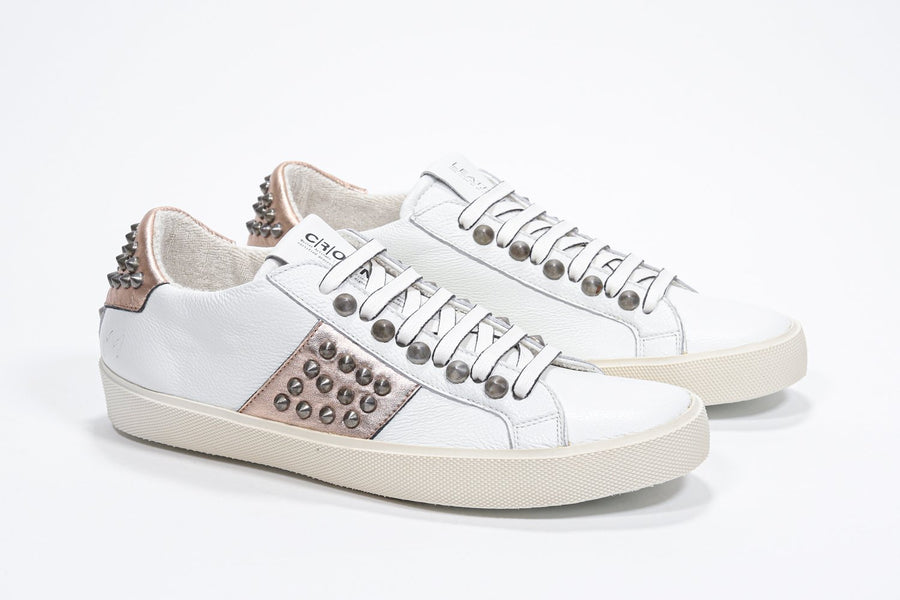 Three quarter front view of low top white and metallic rose sneaker. Full leather upper with studs and vintage rubber sole.