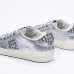 Three quarter back view of low top metallic silver sneaker. Full leather upper with studs and vintage rubber sole.