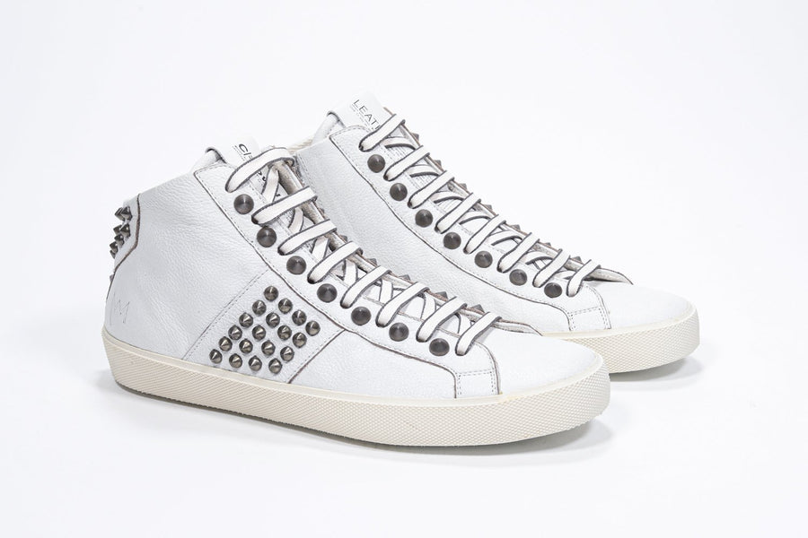 Three quarter front view of mid top white sneaker. Full leather upper with studs, an internal zip and vintage rubber sole.