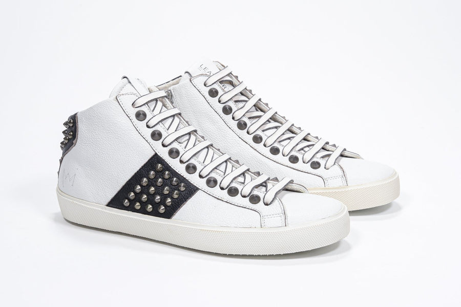 Three quarter front view of mid top white and black sneaker. Full leather upper with studs, an internal zip and vintage rubber sole.