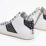 Three quarter back view of mid top white and black sneaker. Full leather upper with studs, an internal zip and vintage rubber sole.