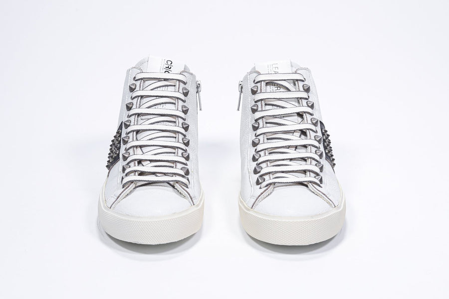 Front view of mid top white and black sneaker. Full leather upper with studs, an internal zip and vintage rubber sole.