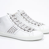 Three quarter front view of mid top white sneaker. Full leather upper with studs, an internal zip and white rubber sole.