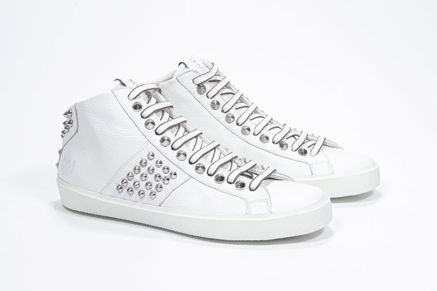 Three quarter front view of mid top white sneaker. Full leather upper with studs, an internal zip and white rubber sole.