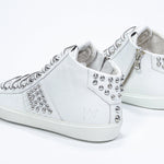 Three quarter back view of mid top white sneaker. Full leather upper with studs, an internal zip and white rubber sole.