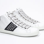 Three quarter front view of mid top white and black sneaker. Full leather upper with studs, an internal zip and white rubber sole.