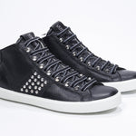 Vista frontale di tre quarti del modello mid top nero sneaker. Tomaia in pelle con borchie, zip interna e suola in gomma bianca.