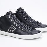 Three quarter front view of mid top black sneaker. Full leather upper with studs, an internal zip and white rubber sole.