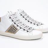 Three quarter front view of mid top white and metallic gold sneaker. Full leather upper with studs, an internal zip and vintage rubber sole.