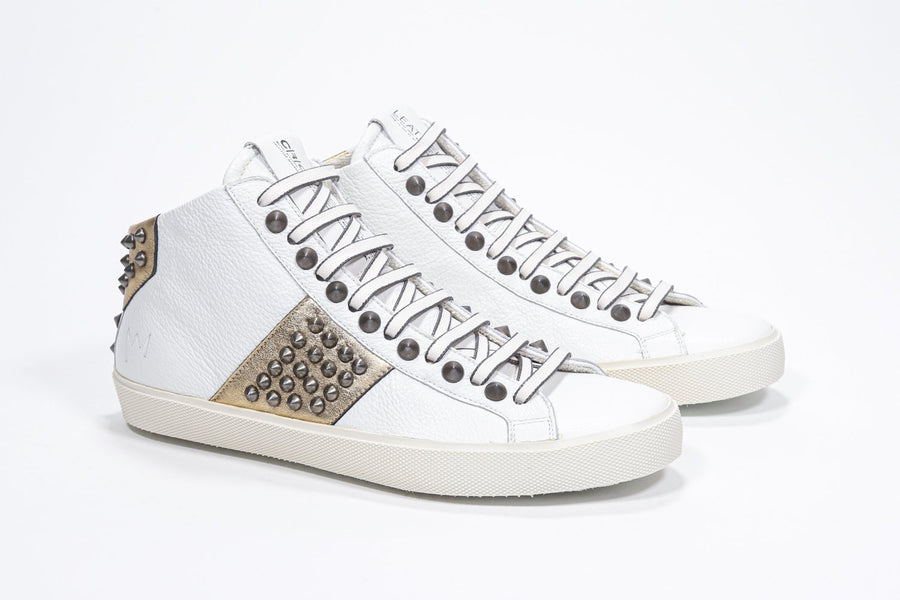 Three quarter front view of mid top white and metallic gold sneaker. Full leather upper with studs, an internal zip and vintage rubber sole.