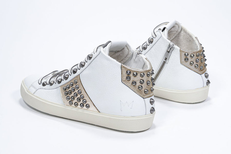 Three quarter back view of mid top white and metallic gold sneaker. Full leather upper with studs, an internal zip and vintage rubber sole.