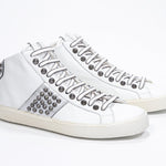 Three quarter front view of mid top white and metallic silver sneaker. Full leather upper with studs, an internal zip and vintage rubber sole.