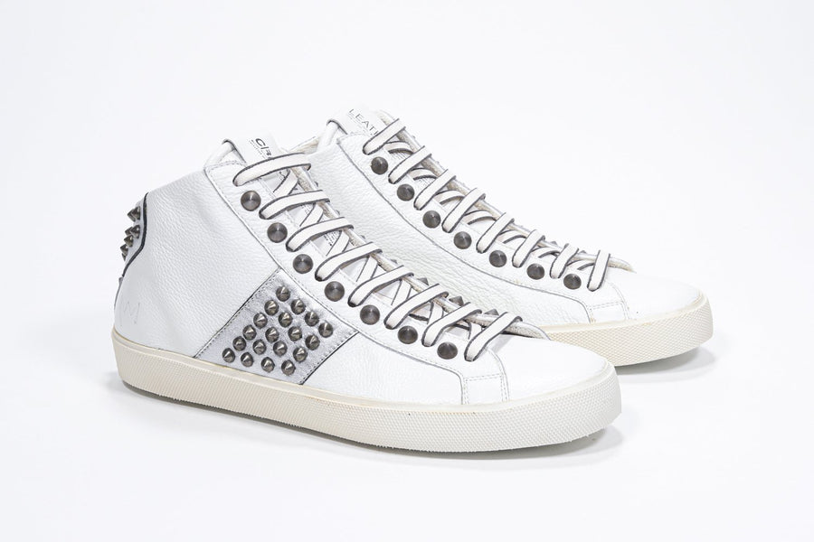Three quarter front view of mid top white and metallic silver sneaker. Full leather upper with studs, an internal zip and vintage rubber sole.
