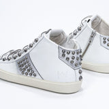 Three quarter back view of mid top white and metallic silver sneaker. Full leather upper with studs, an internal zip and vintage rubber sole.