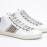 Three quarter front view of mid top white and metallic rose sneaker. Full leather upper with studs, an internal zip and vintage rubber sole.