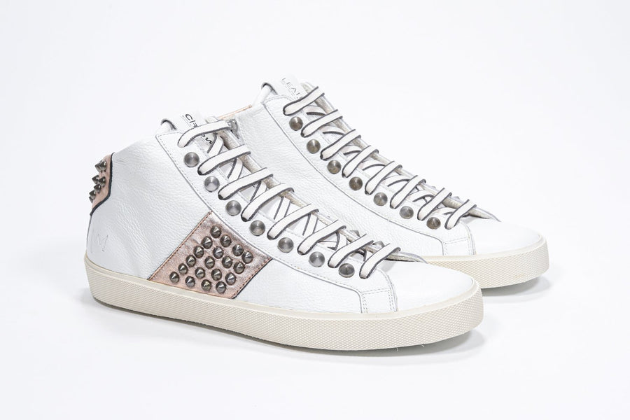 Three quarter front view of mid top white and metallic rose sneaker. Full leather upper with studs, an internal zip and vintage rubber sole.