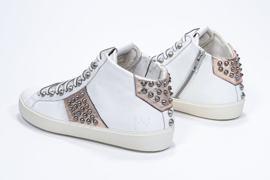 Three quarter back view of mid top white and metallic rose sneaker. Full leather upper with studs, an internal zip and vintage rubber sole.